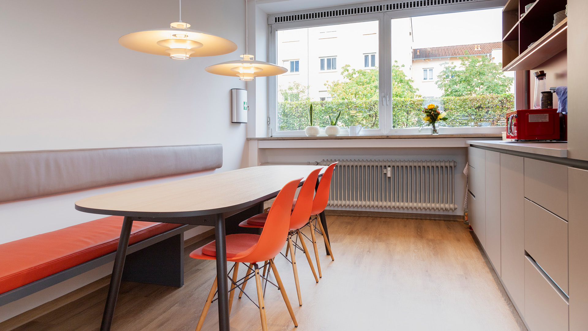 Aufenthaltsbereich mit Küche mit Sitzbank, langem Holztisch und orangefarbenen Stühlen.
