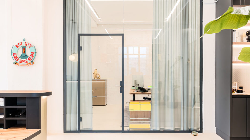 Glastrennwand mit Vorhängen zur flexiblen Zonierung im Referenzprojekt Heimathafen Immobilien in Münster, mit abtrennbarem Büroarbeitsplatz, Sichtschutz und offener Teeküche im Hintergrund.