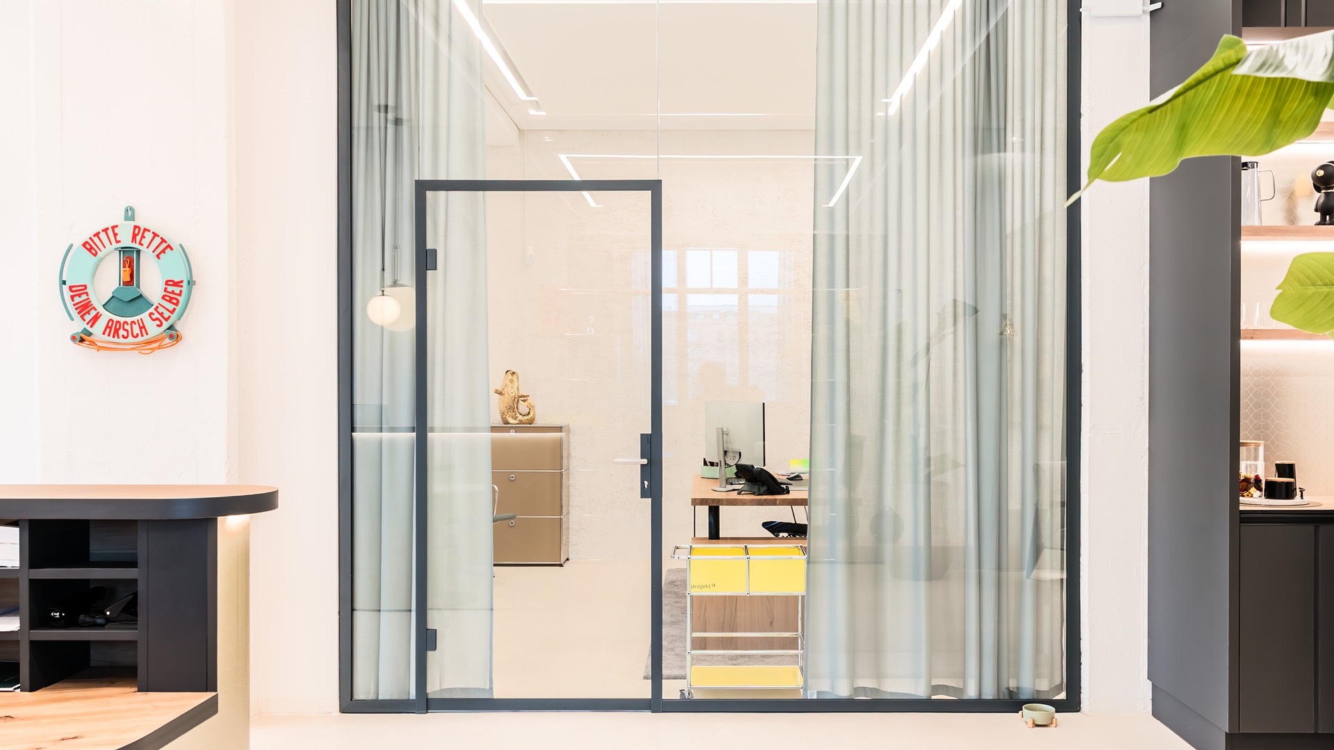Glastrennwand mit Vorhängen zur flexiblen Zonierung im Referenzprojekt Heimathafen Immobilien in Münster, mit abtrennbarem Büroarbeitsplatz, Sichtschutz und offener Teeküche im Hintergrund.